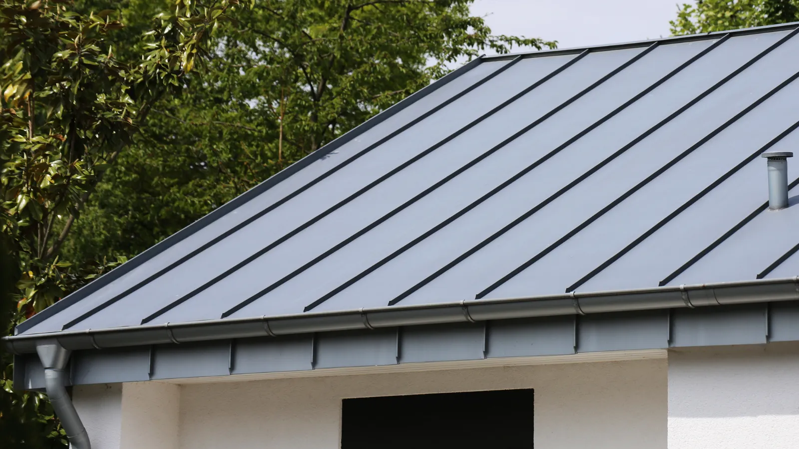 Vent pipe on a gray standing seam metal roof near trees
